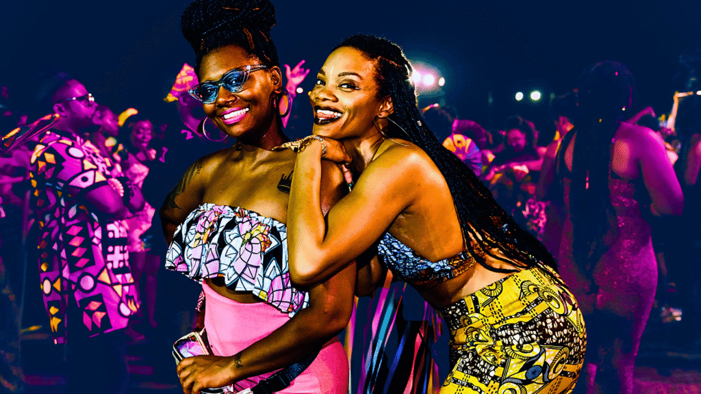 Two women smiling and hugging at a lively nighttime event, dressed in colorful traditional outfits.