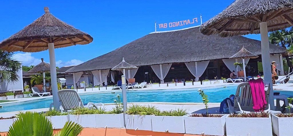 Outdoor view of Flamingo Bay resort near Luanda, Angola, featuring a tropical pool area with thatched umbrellas, sun loungers, palm trees, and a relaxing atmosphere under a bright blue sky.