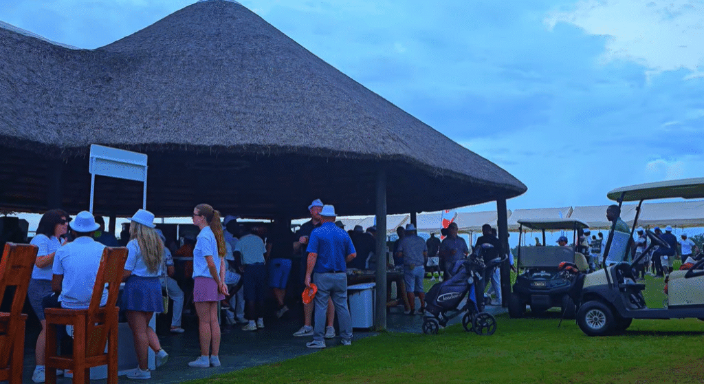 Golfers and guests gathered under a thatched-roof pavilion at Mangais Golf Resort in Angola, surrounded by golf carts, gear, and a green open landscape during a social event.