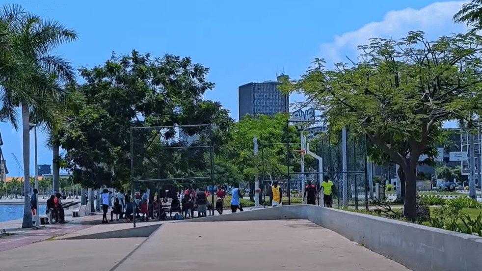 Group of people playing and gathering near a basketball court along the Marginal in Luanda, Angola, surrounded by palm trees, city buildings and open pedestrian spaces under a bright blue sky.