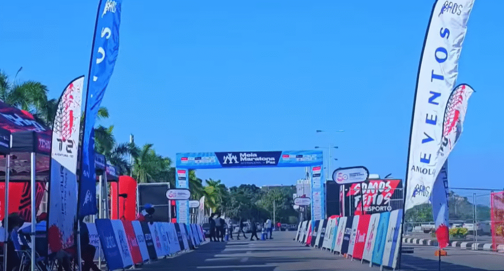 Start or finish line of the Corrida da Paz (Peace Race) on Luanda’s New Marginal, featuring colourful flags, sponsor banners, palm trees and event structures under a clear blue sky.