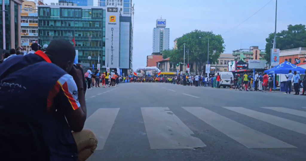 Street-level view of the São Silvestre race in Luanda, Angola, with a crowd lining the city avenue, a photographer capturing the scene, and corporate buildings in the background.