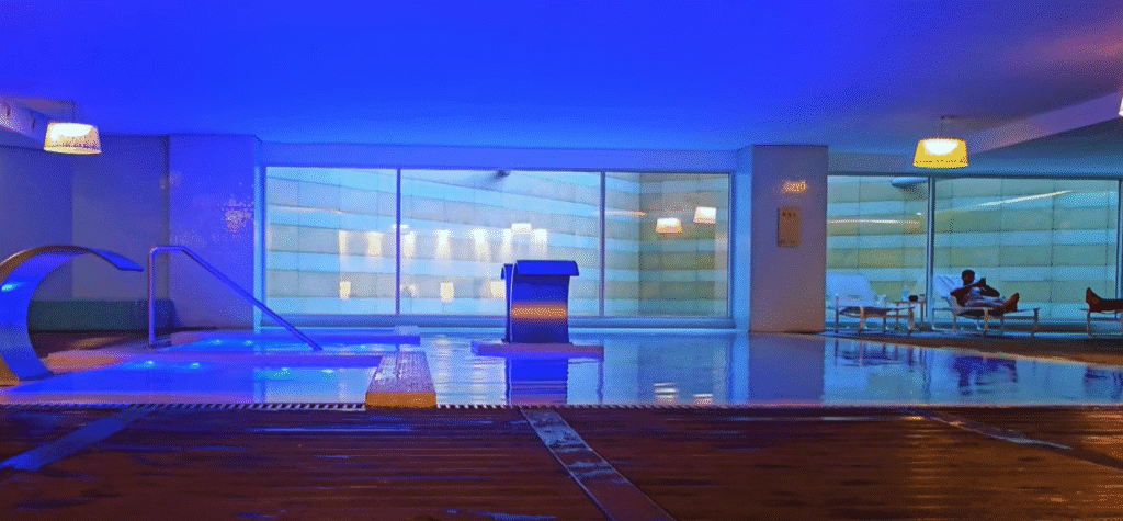 Indoor spa pool at Epic Sana Luanda, featuring modern lighting, wooden flooring, stainless steel water jets, and a tranquil atmosphere with guests relaxing on loungers.