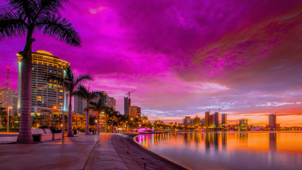 Luanda waterfront skyline at sunset with palm trees and modern buildings.