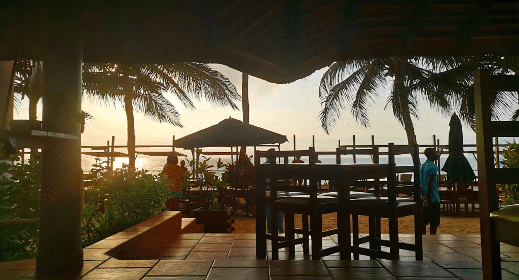 Sunset view from inside Boutique Lodge Sangano in Angola, overlooking palm trees, beach seating, and the ocean. The warm golden light and tropical setting create a peaceful and inviting retreat atmosphere.