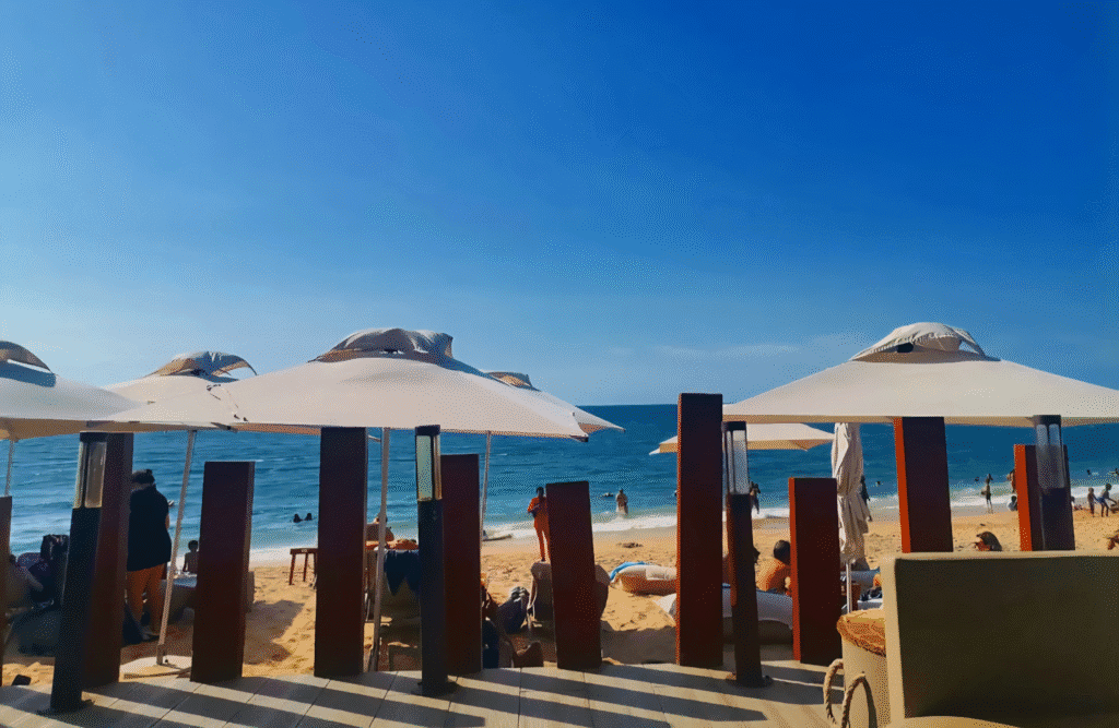 Café del Mar Luanda – beachside lounge with large white umbrellas, sunbeds, and guests relaxing by the sea under a clear blue sky.