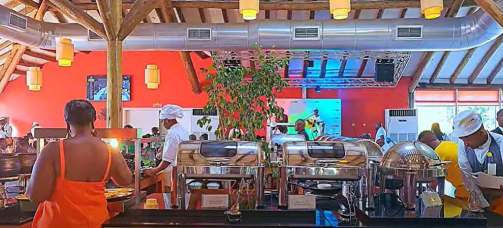 A vibrant buffet restaurant in Luanda with chefs serving guests, colorful decor, and a lively atmosphere. A woman in an orange dress selects food, while others enjoy the meal in a modern, warmly lit space with wooden beams and industrial ventilation.