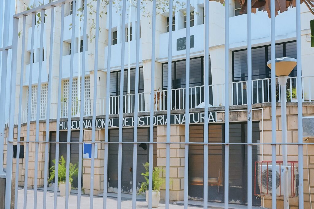 A photograph of the exterior of the National Museum of Natural History in Luanda, Angola, showing its three-story building behind protective metal bars. The image highlights the museum’s historic architecture and urban setting in the Ingombota district.