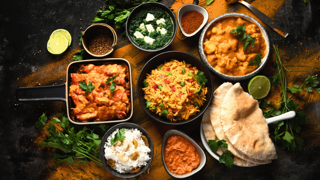 Top view of a traditional Indian meal featuring various curries, spiced rice, palak paneer, naan bread, white rice with raisins and cashews, and chutney, all arranged on a dark surface with scattered spices and fresh herbs.