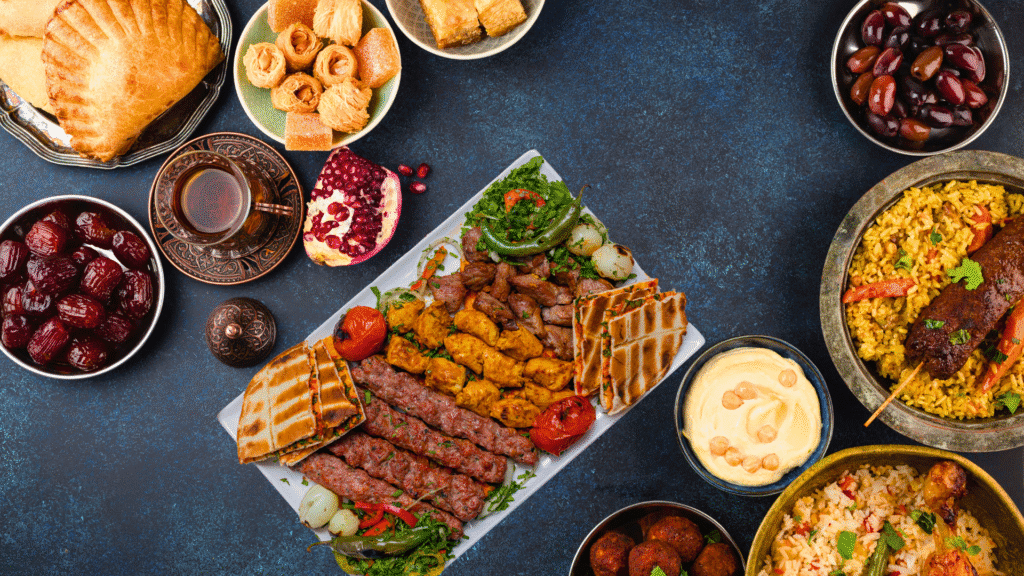 Middle Eastern feast featuring grilled kebabs, chicken skewers, lamb kofta, flatbreads, hummus, seasoned rice dishes, olives, dates, pastries like baklava and ma'amoul, a pomegranate, and a traditional glass of tea, all arranged on a blue background.