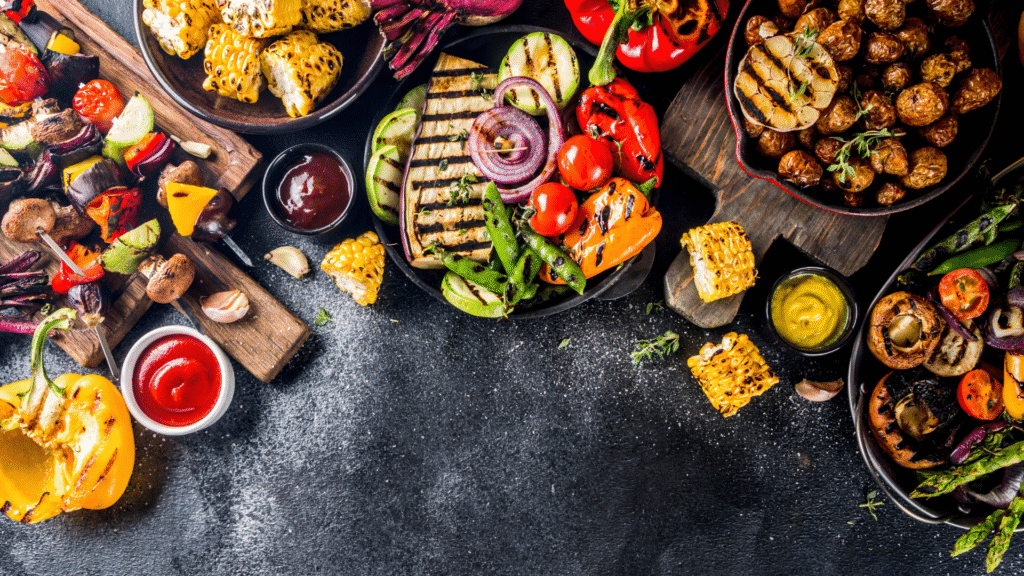 Colorful assortment of grilled vegetables including bell peppers, corn on the cob, zucchini, mushrooms, onions, potatoes, and eggplant, served with dipping sauces on a dark rustic background, showcasing a vibrant vegetarian barbecue spread.