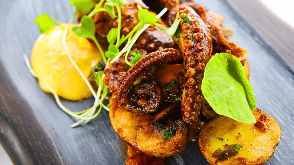 Portuguese-style grilled octopus served with roasted potatoes, fresh herbs, and a smooth yellow sauce, beautifully plated on a dark ceramic dish.