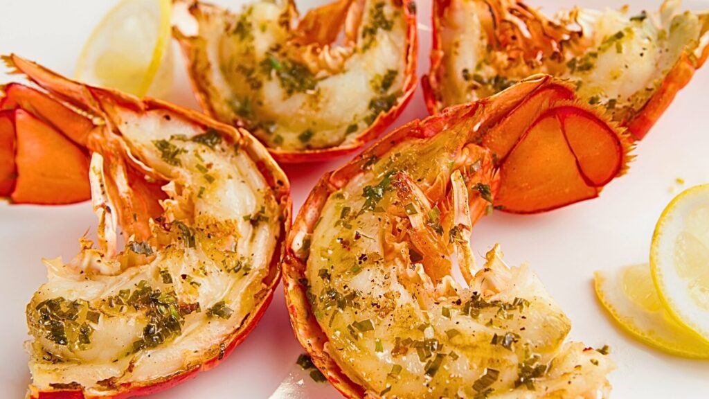 Grilled lobster tails seasoned with garlic, herbs, and olive oil, served with lemon slices on a white plate.