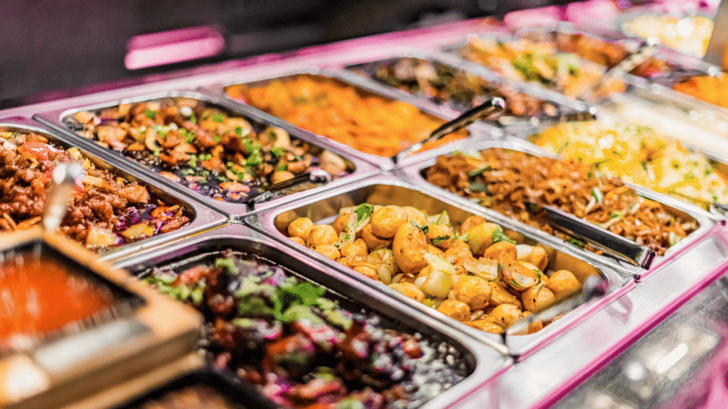 Colorful buffet with various dishes including stir-fried vegetables, noodles, potatoes, and meat dishes in stainless steel trays, ready to be served.