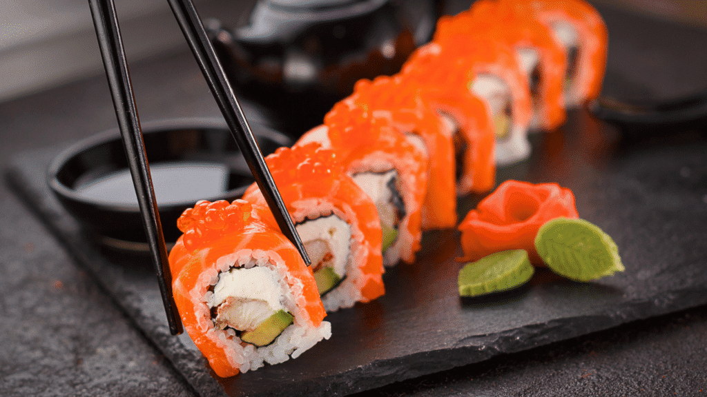 Close-up of sushi rolls topped with fresh salmon and salmon roe, filled with cream cheese, avocado, and crab, served on a black slate plate with soy sauce and decorative wasabi and ginger shaped like a rose.