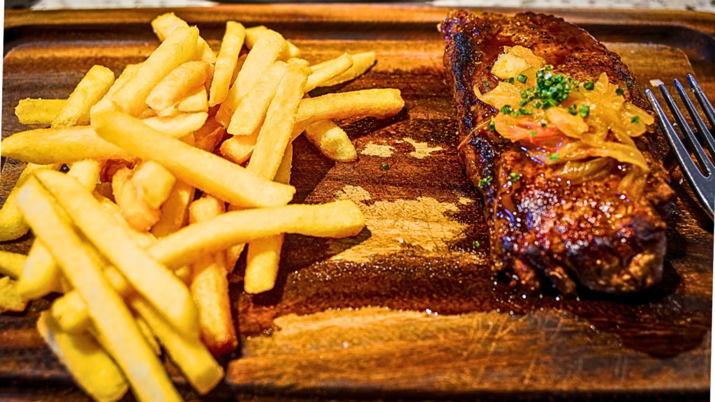 Portuguese-style grilled steak topped with sautéed onions, served with golden French fries on a rustic wooden board.