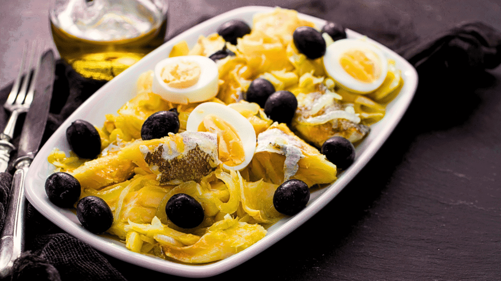 Traditional Portuguese dish 'Bacalhau à Gomes de Sá' with flaked salted codfish, sliced boiled eggs, black olives, onions, and potatoes, served on a white plate.