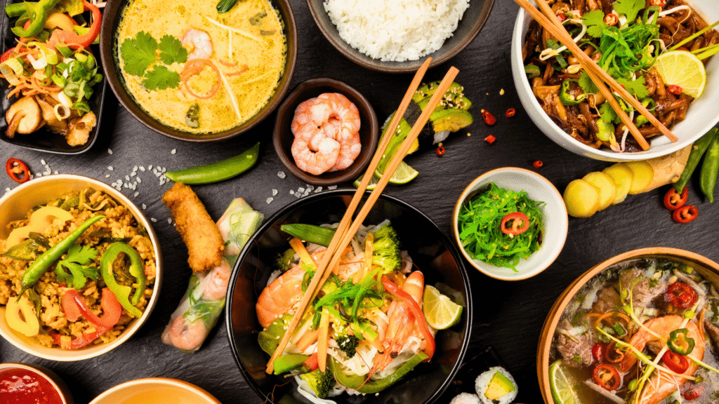 Top view of a colorful Asian food assortment including noodle soup with shrimp, fried rice, spring rolls, green curry, sushi, stir-fried noodles, and various fresh vegetables and herbs, arranged on a dark slate surface with chopsticks.