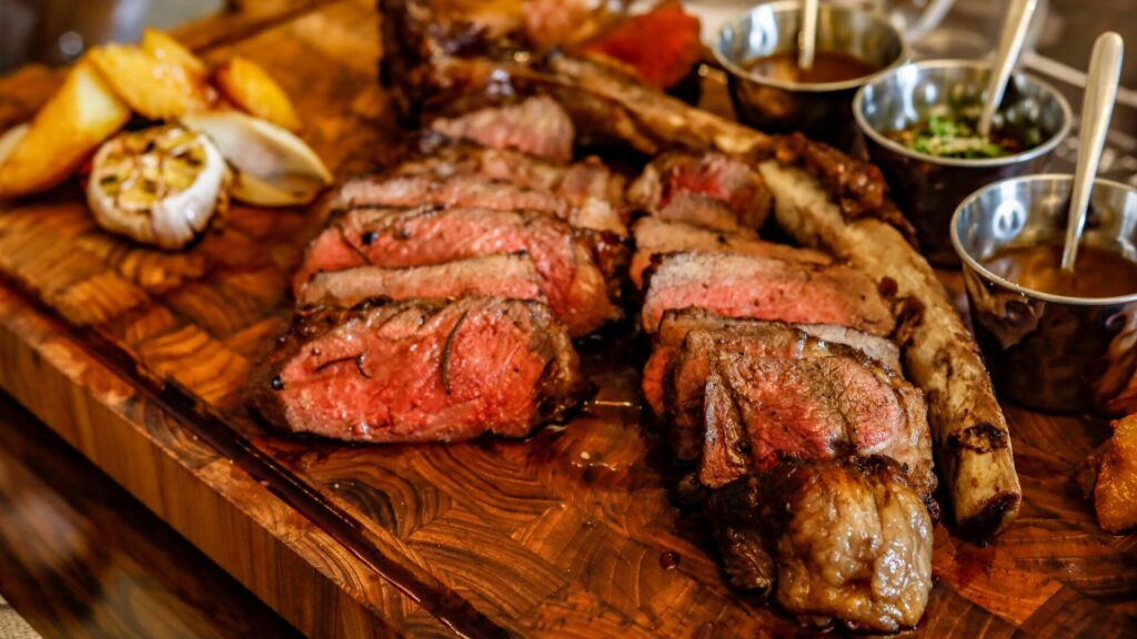 Juicy grilled ribeye steak sliced on a wooden board, served with roasted potatoes, garlic, and a variety of sauces.