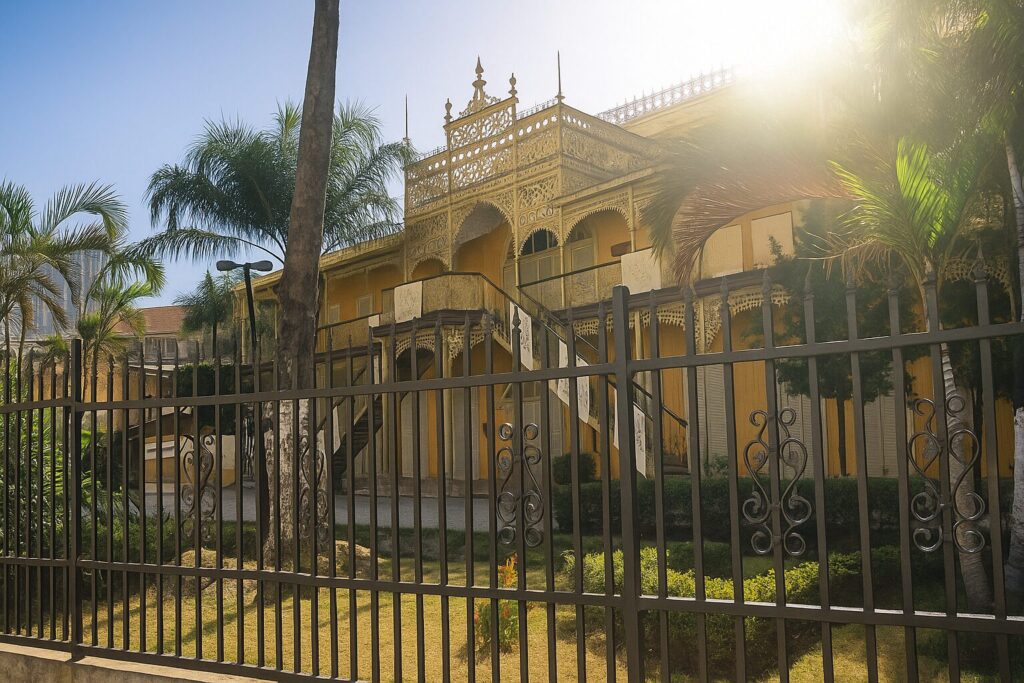 A digital photograph of the Iron Palace (Palácio de Ferro) in Luanda, Angola, showing its ornate ironwork facade, tropical garden, and historic yellow building behind a decorative metal fence on a sunny day.