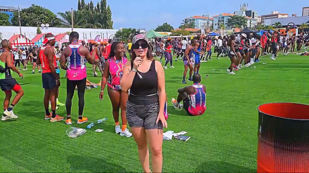 A woman speaking into a microphone during the Talatona Fitness Fair 2025, surrounded by athletes and participants on a vibrant green field, with people dancing and exercising in the background.