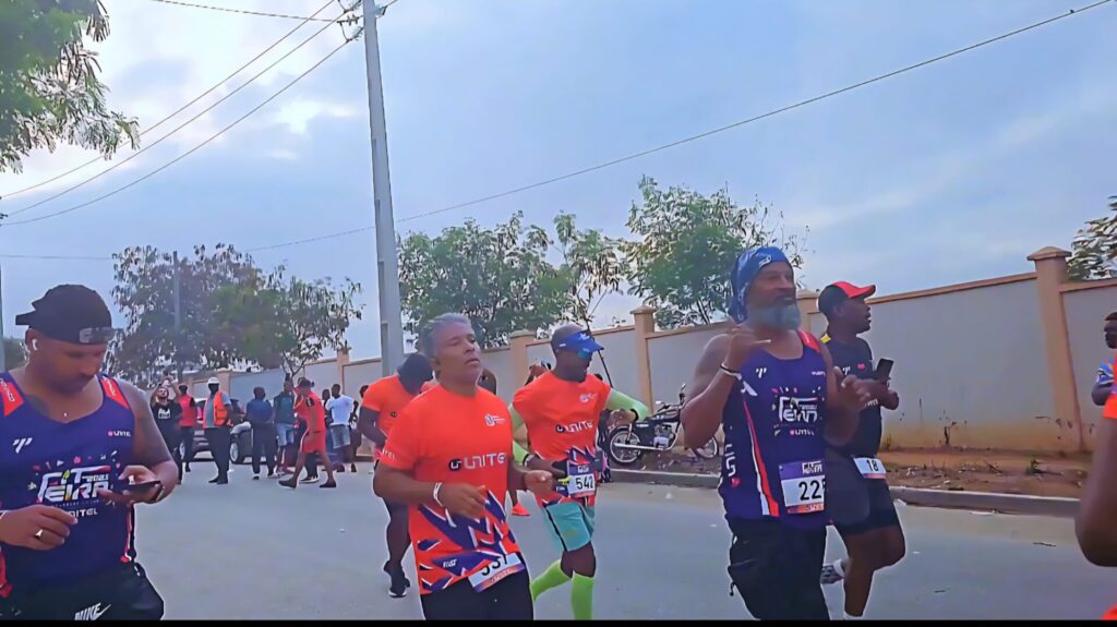 Participants running during the early morning race at the Talatona Fitness Fair 2025, wearing event shirts and bib numbers, surrounded by fellow runners and onlookers.