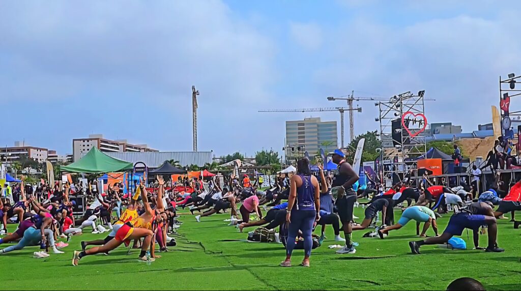 Dozens of participants stretching together during a large outdoor fitness session at the Talatona Fitness Fair 2025, with tents, booths and Luanda’s skyline in the background.