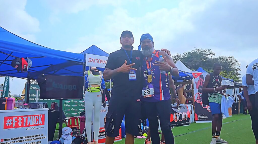 Two men posing in front of the FitNick booth at the Talatona Fitness Fair 2025, one wearing a race medal and the other representing the brand, with sports apparel and promotional banners around them.