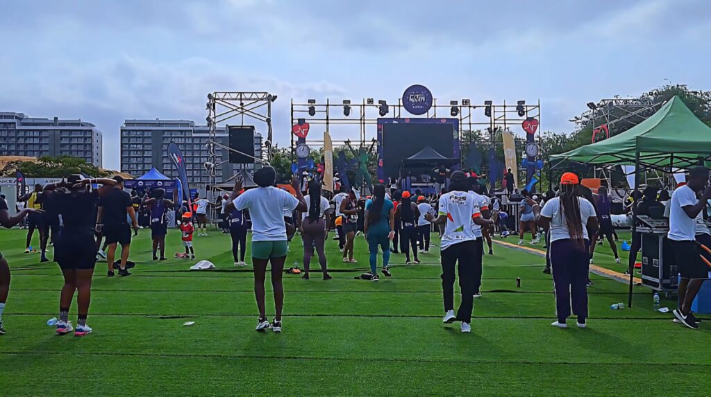 Group fitness activity in front of a main stage at the Talatona Fitness Fair 2025, with instructors leading the crowd and tall buildings in the distance.