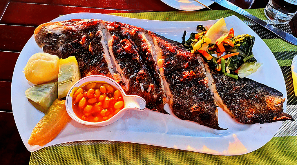 Traditional Angolan grilled fish served with cassava, sweet potato, plantain, beans in sauce, and sautéed vegetables on a white plate.